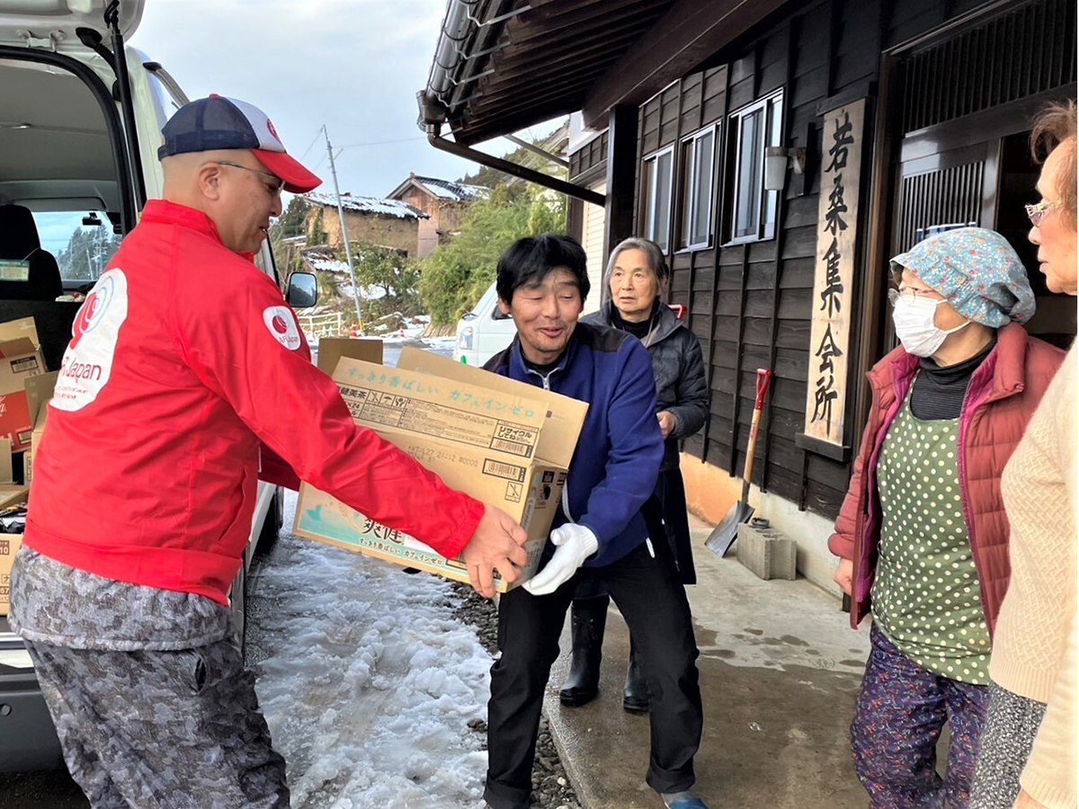 段ボールを手渡すAAR職員とそれを受け取る地元の男性　それを見守る地元の人々