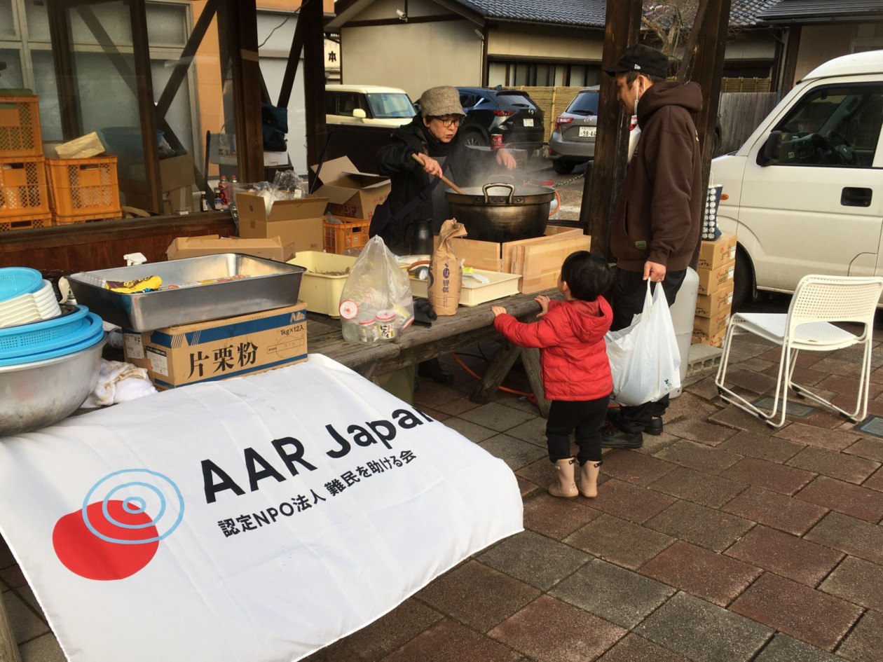 炊き出しに並ぶ親子と鍋から汁物をつぐスタッフ。手前はAAR Japanのバナー