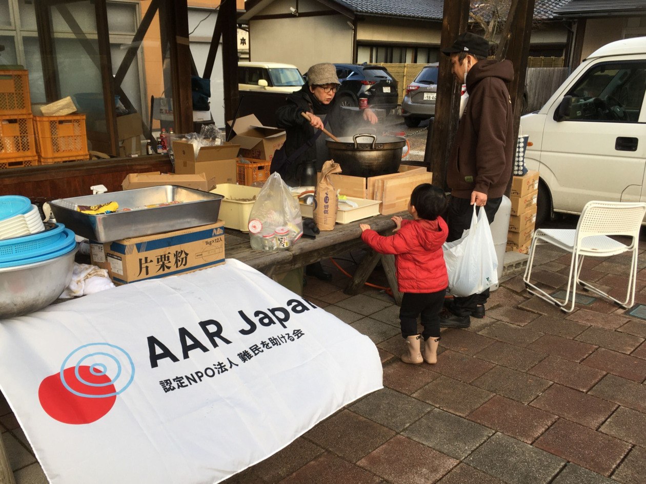 A parent and a child lined up for the soup kitchen, and staff pouring soup from the pot. AAR Japan banner in the foreground.