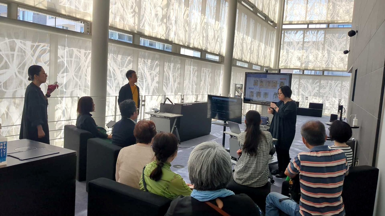 手話による説明と同時に、手話通訳とAI音声認識による字幕表示も用いた事前説明会の様子。説明者が二つのモニター(メインと字幕用)の横に立って手話で説明。字幕用モニターには手話通訳士が読み取り翻訳した音声をAI認識し、字幕化して表示している。